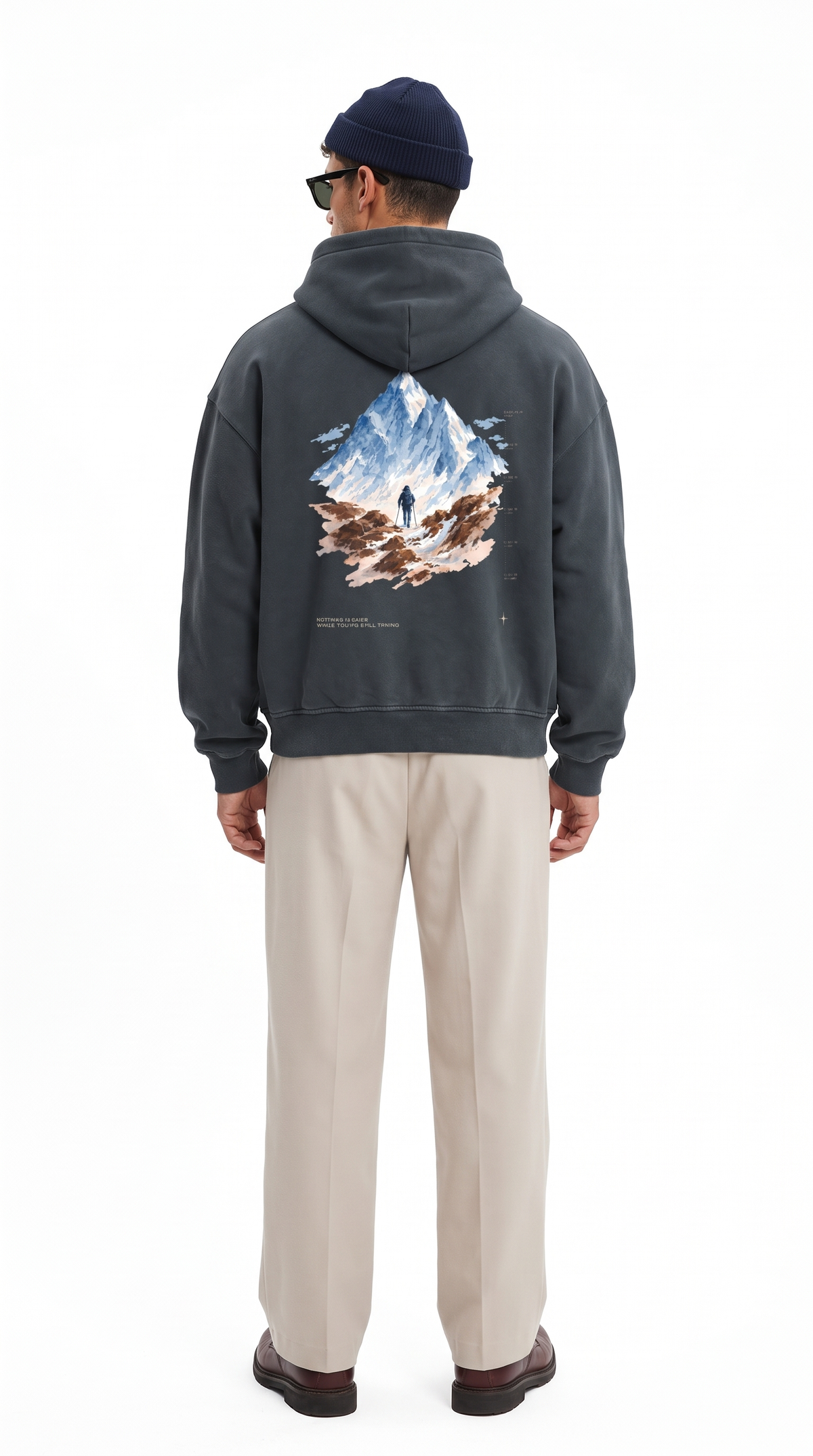 A man viewed from the back wearing a dark charcoal gray hoodie with a large mountain graphic on the back, paired with beige trousers, a navy beanie, and sunglasses, against a plain white background.