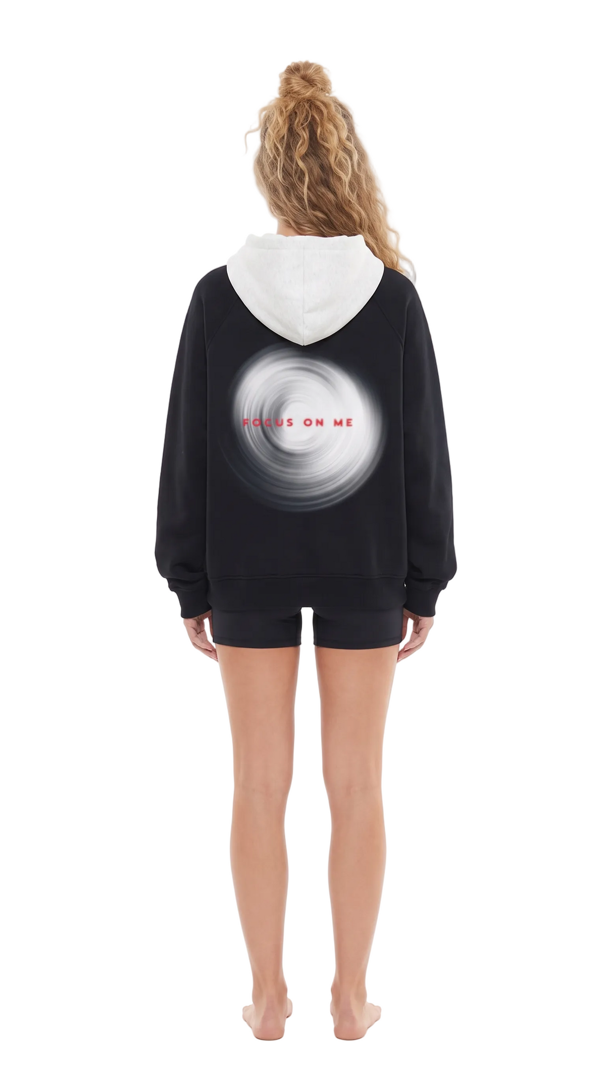 A woman viewed from behind wearing a black hoodie with a white hood and a large white circular graphic on the back featuring the red text 'FOCUS ON ME'. She is also wearing black shorts against a plain white background.