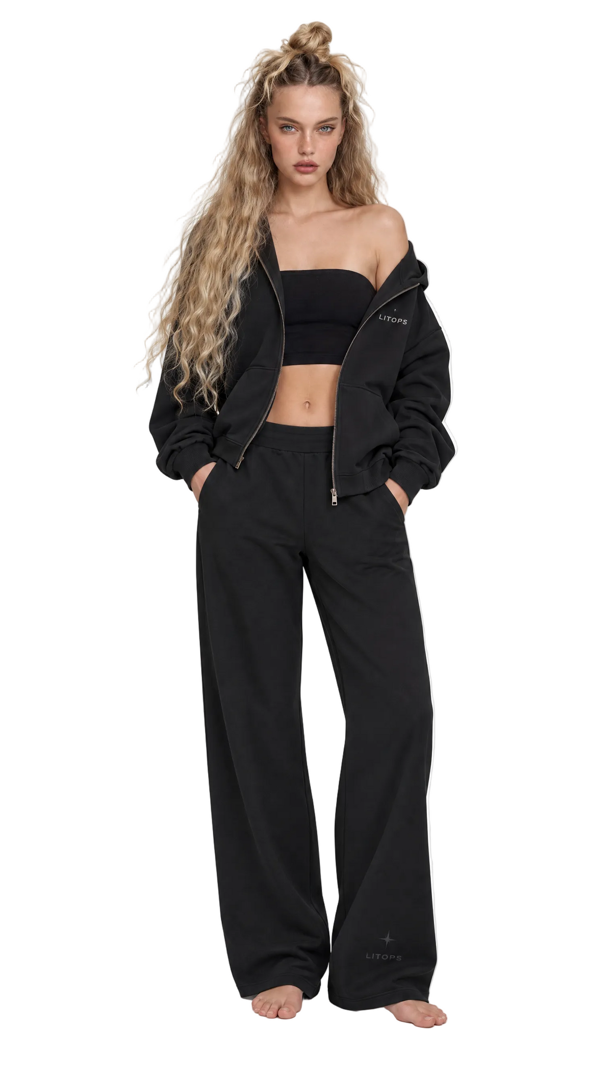 A woman modeling a black lounge set consisting of a cropped zip-up hoodie and matching wide-leg sweatpants, styled with a black strapless bandeau top against a plain white background.
